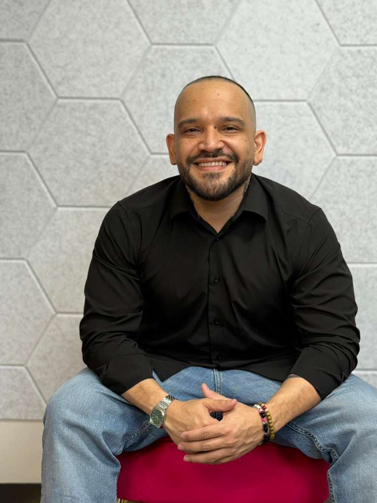 Smiling man with a beard, wearing a black shirt and jeans, sitting on a pink cushion against a hexagonal wall.