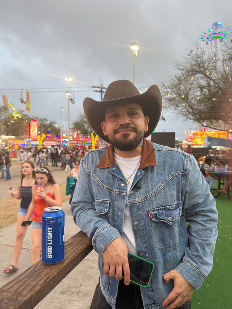 Man wearing a cowboy hat and denim jacket at fairground with rides and people in background