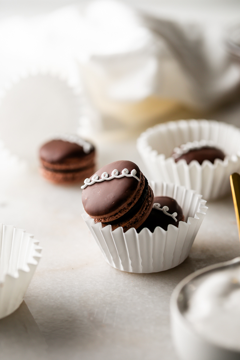 Chocolate macarons in white paper cups on a light surface, with blurred background.