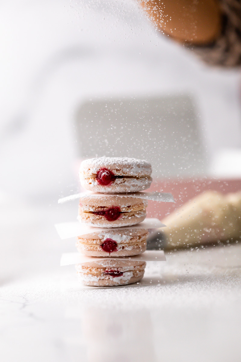 Stack of four cookies with red berries, dusted with powdered sugar, on a white surface with a blurred background.