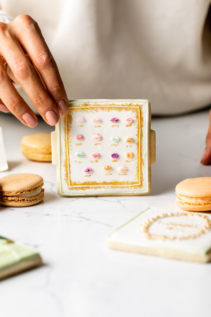 Person's hand holding a decorated cookie with colorful icing, surrounded by macarons on a white surface.