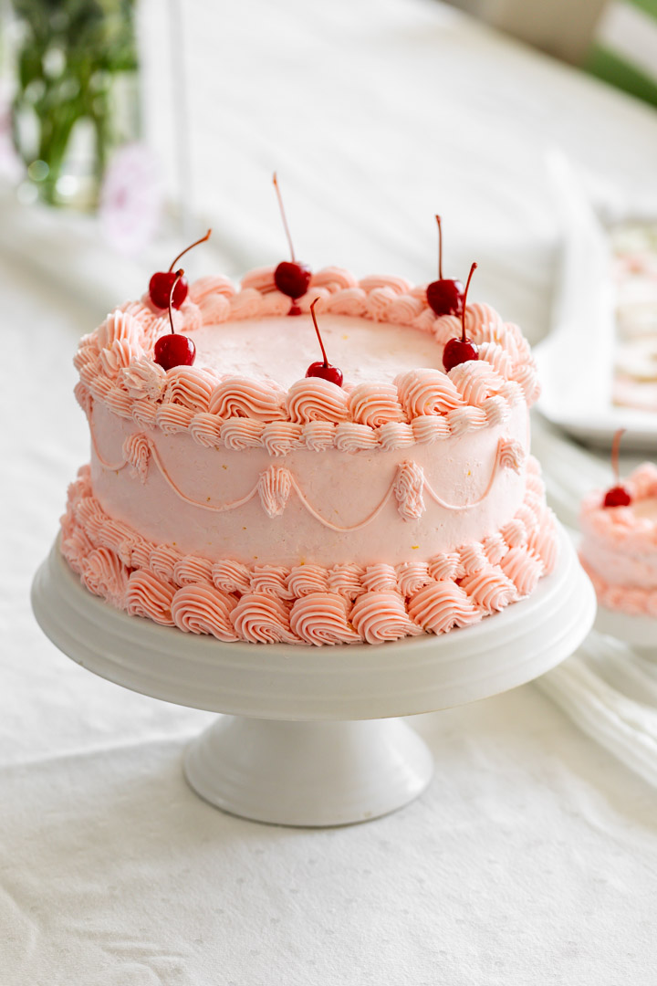 Pink decorated cake with cherries on top on a white cake stand, with a blurred background of a bed and green plant.