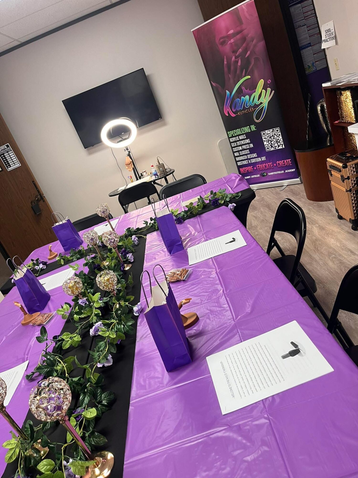 Conference table with purple tablecloth, decorated with flowers and purple gift bags, in a room with a banner and a TV.