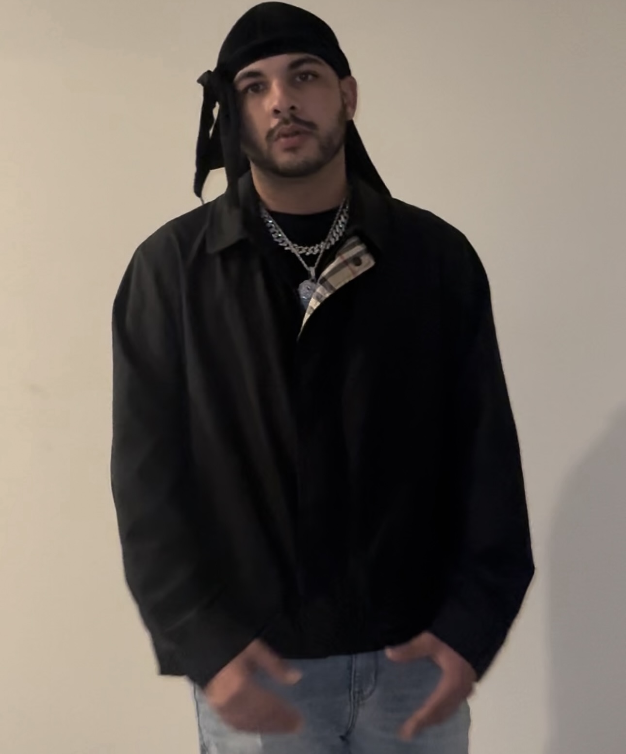Young man with a black headscarf, black jacket, and layered necklaces, standing against a plain wall.