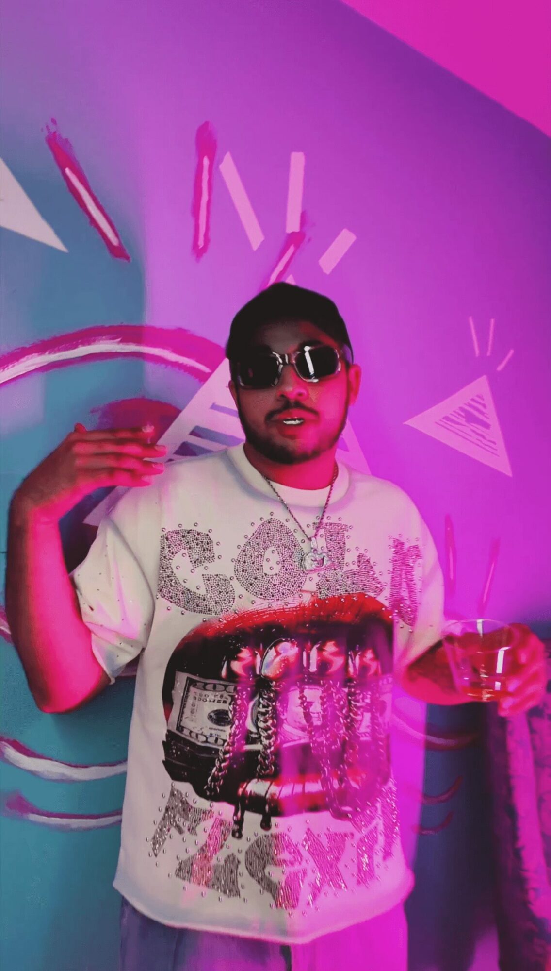 Man wearing sunglasses and a graphic T-shirt, holding a drink, standing in front of a colorful, abstract background.