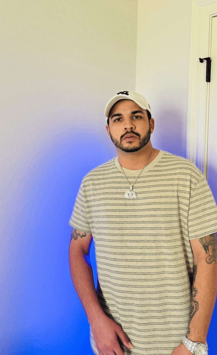 Young man with a beard, wearing a striped t-shirt, white cap, and jewelry, standing indoors with a blue light behind him.
