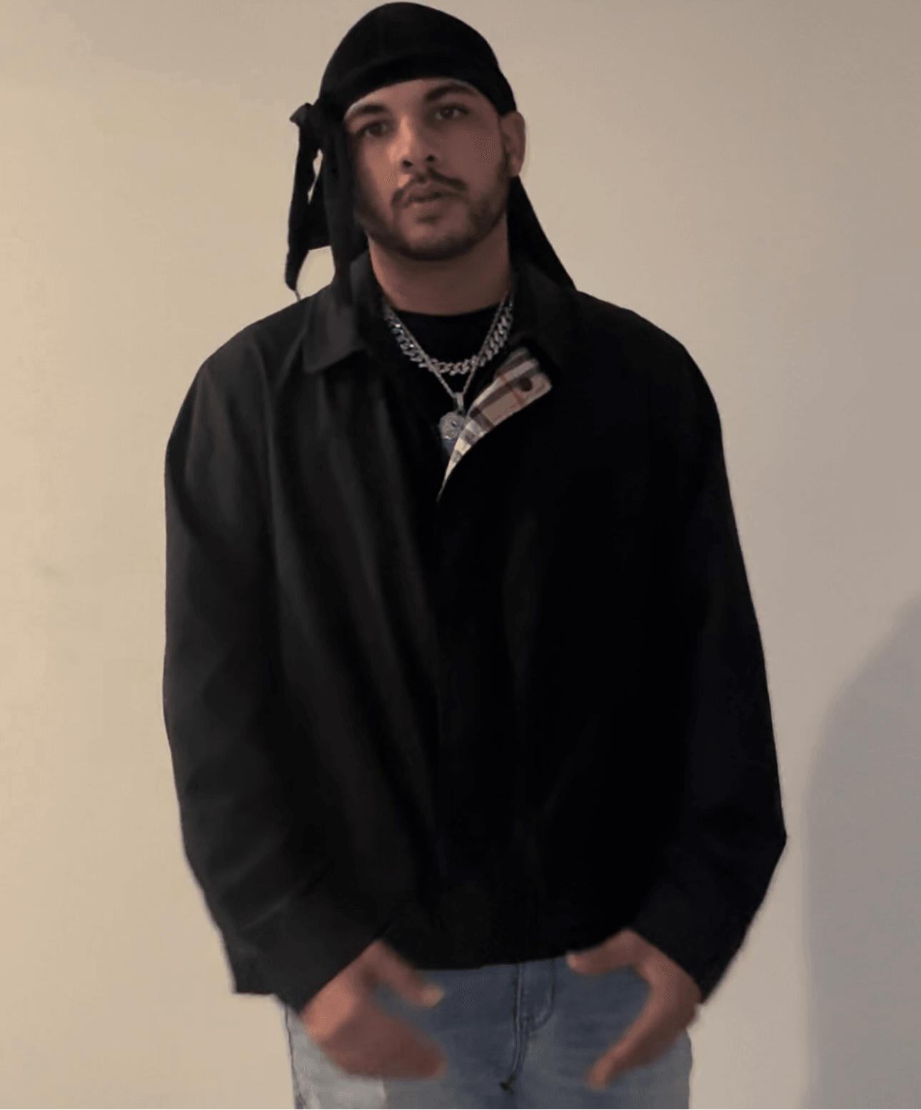 Young man with a black headscarf, black jacket, and layered necklaces, standing against a plain wall.