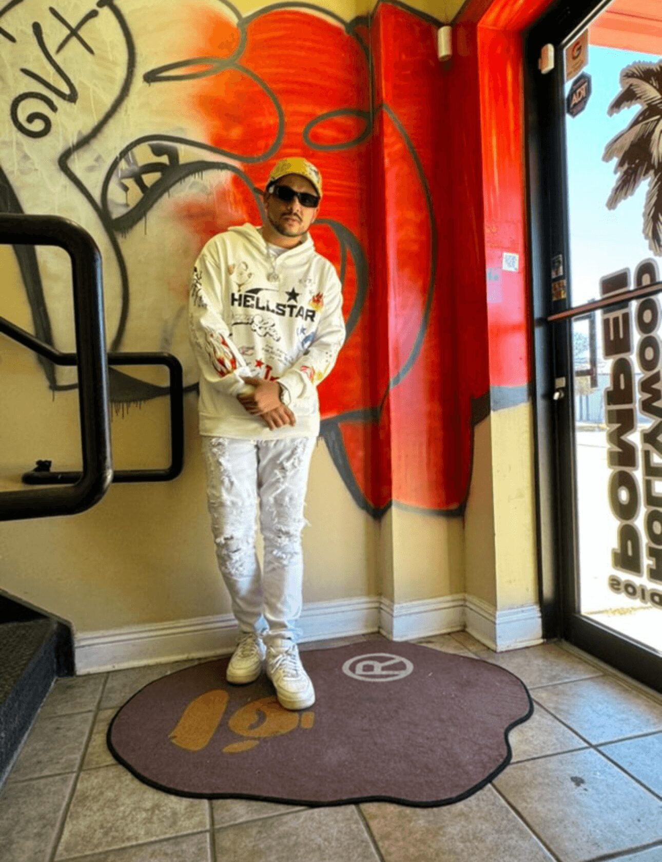 Person wearing sunglasses and a white outfit stands on a brown rug inside a colorful room with graffiti art and a glass door.