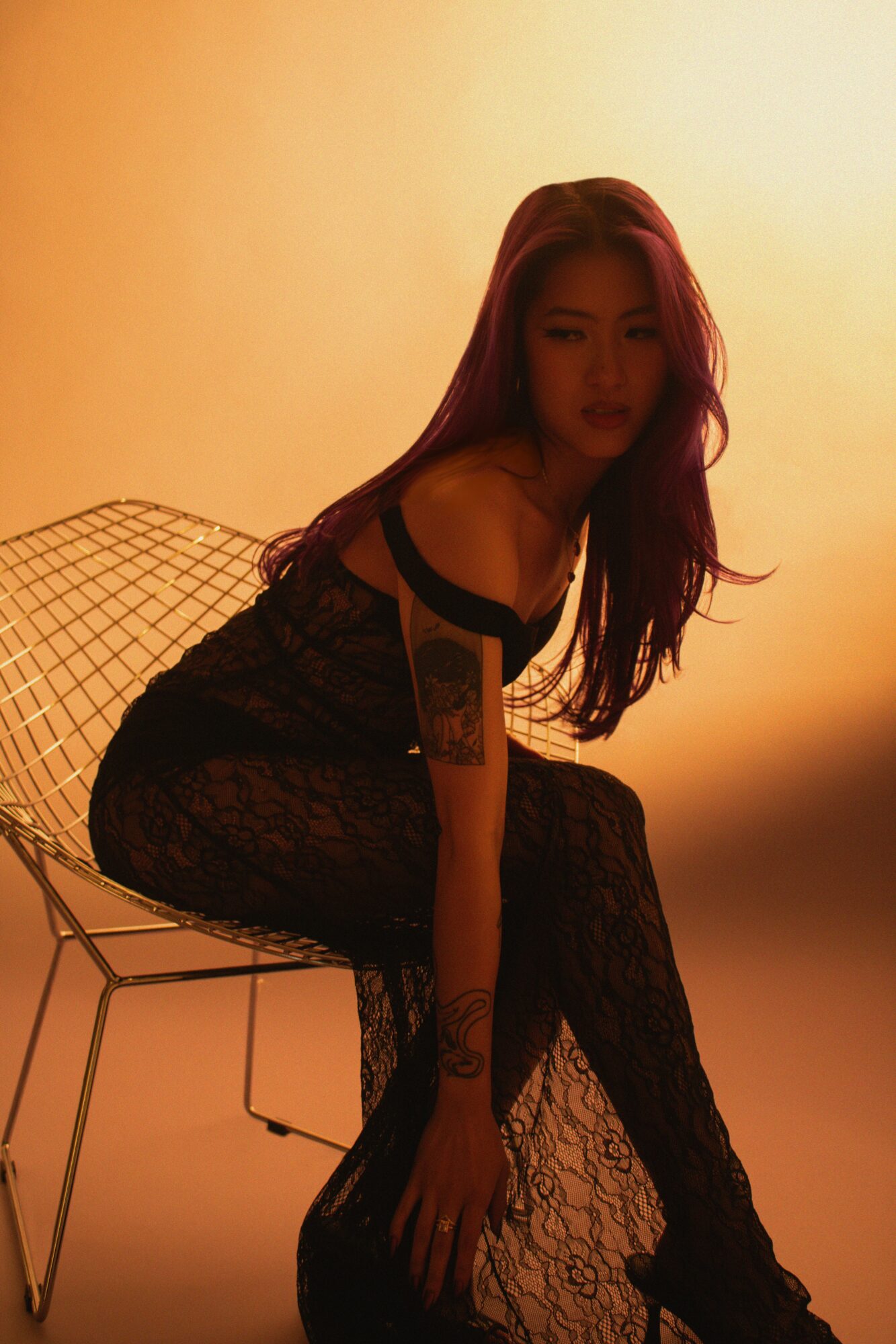 Woman with long hair sitting on a wire chair, wearing a lace dress, in warm lighting.