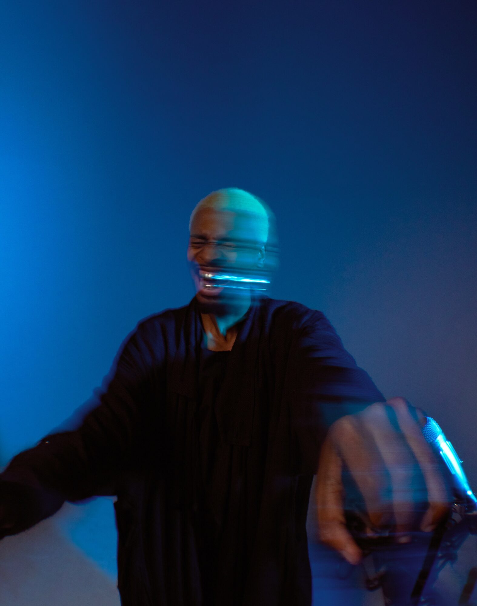 Person with bald head smiling, reaching out with hand, blurred motion, dark background, blue lighting