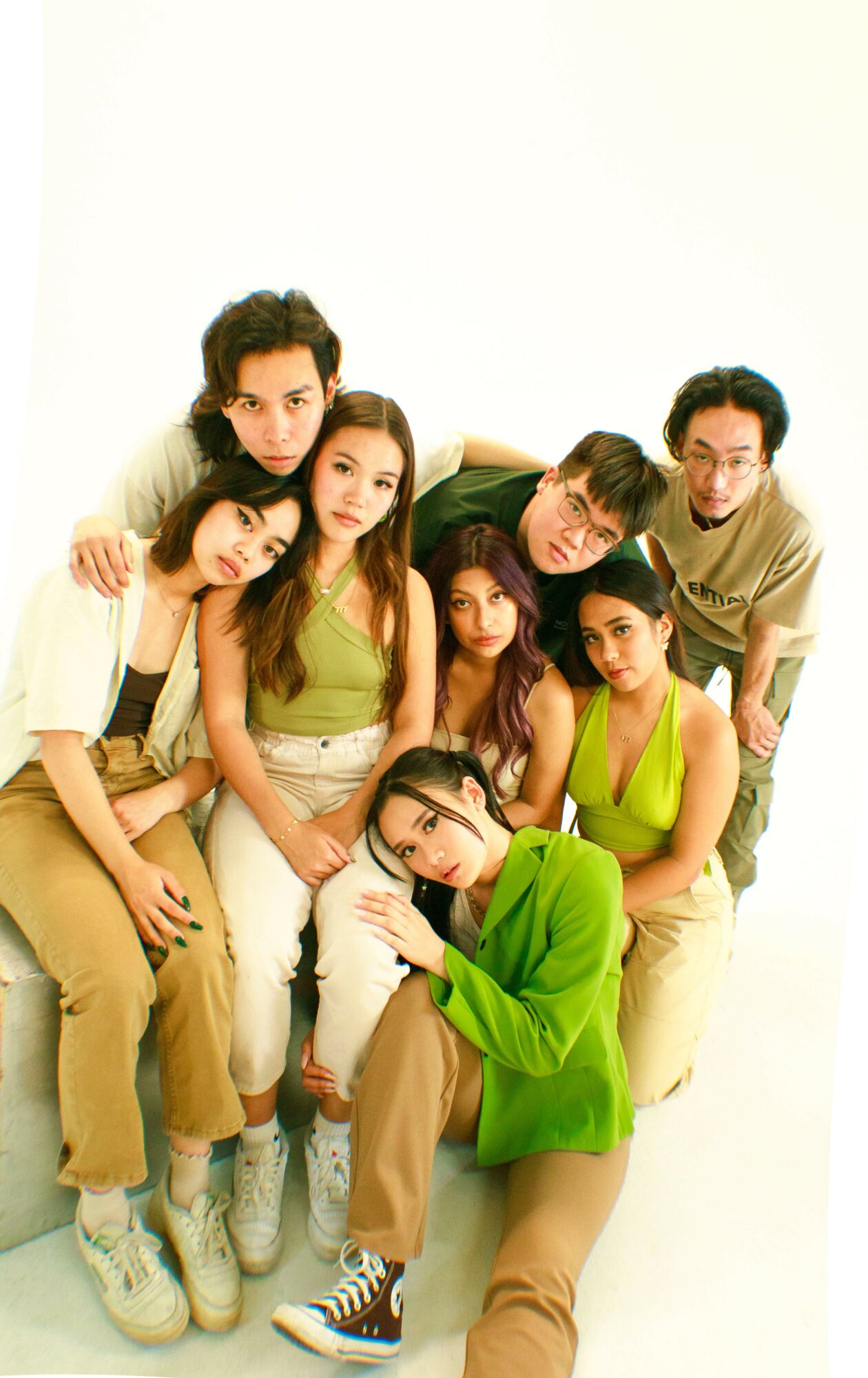Group of seven young people sitting together, some leaning on each other, against a plain white background.