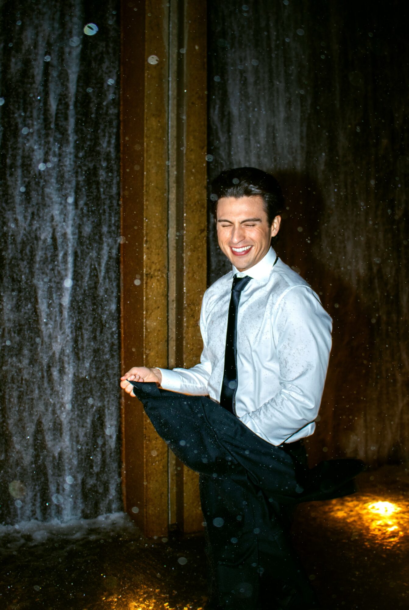 Man in formal attire smiling under a waterfall, holding a jacket, with water splashing around him.