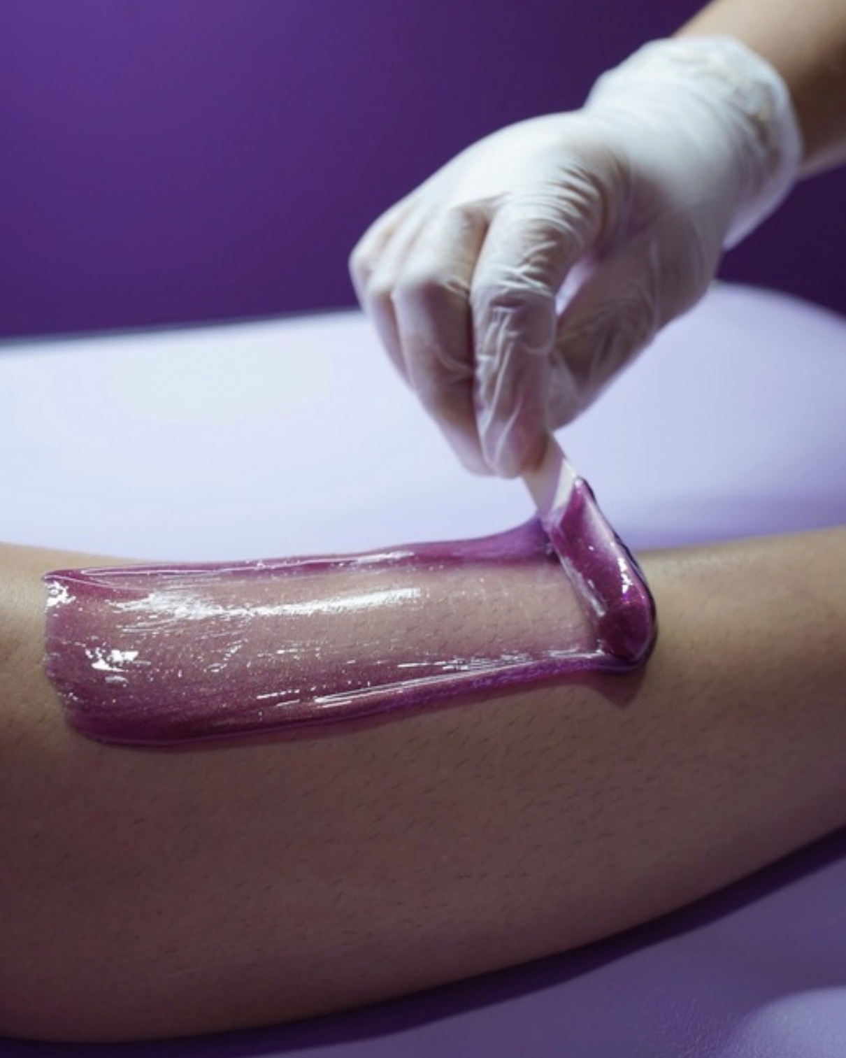 Person applying purple wax to a person's leg during a waxing procedure, wearing a glove.