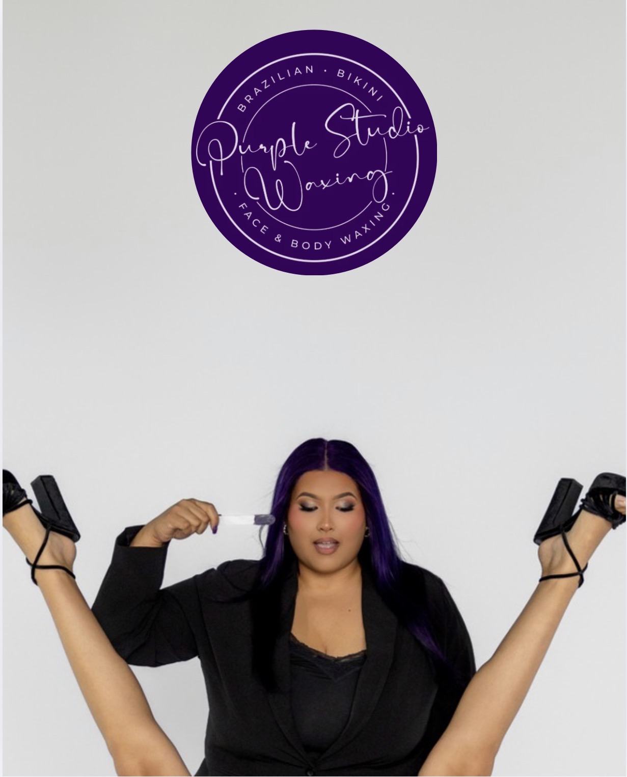Woman with purple hair lying on her back with arms and legs raised, wearing black clothing, in front of a white wall with purple logo.