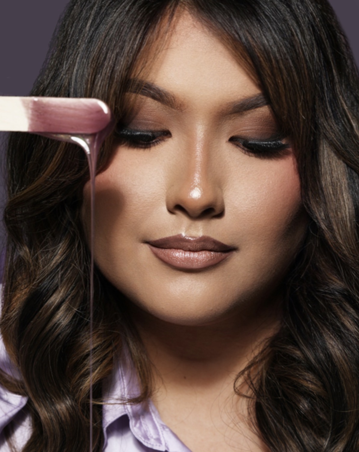Close-up of a woman with dark wavy hair and makeup, holding a makeup brush near her face.