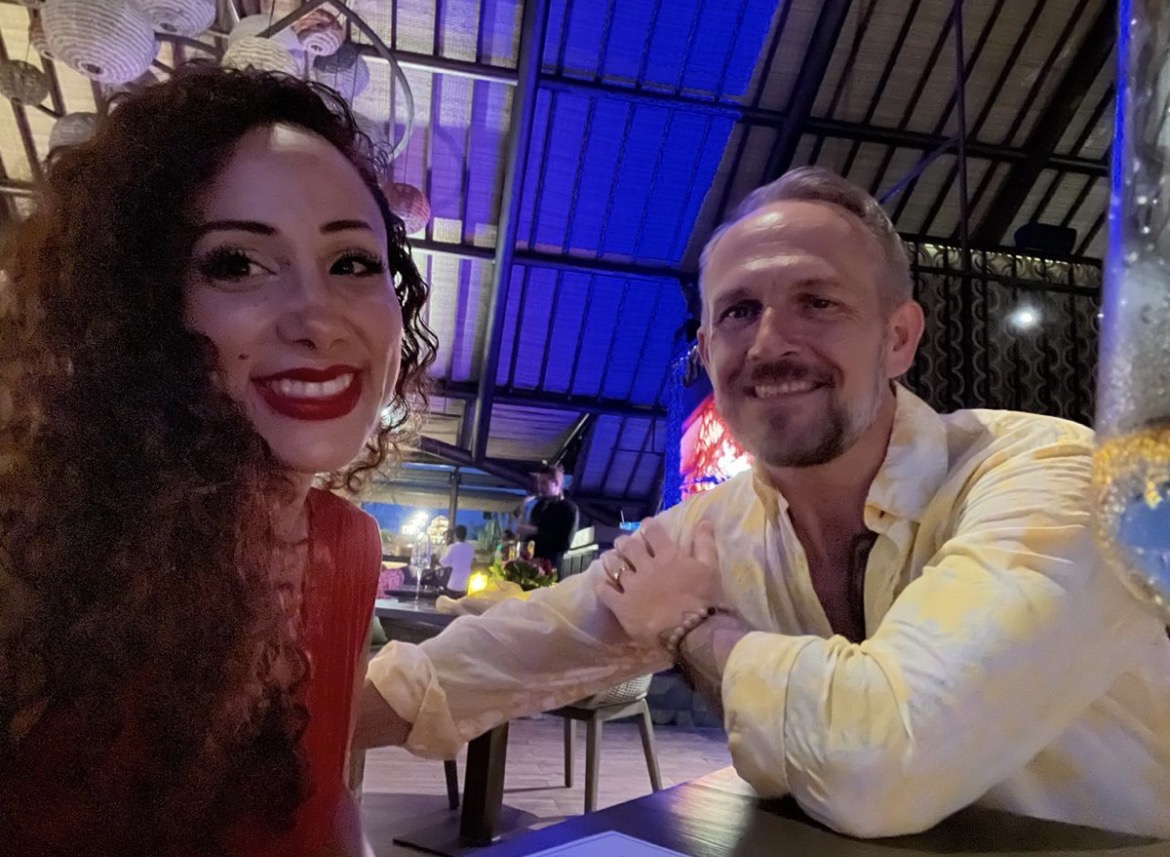 A smiling woman with curly hair and a man with short hair sit at a table in a decorated indoor setting.