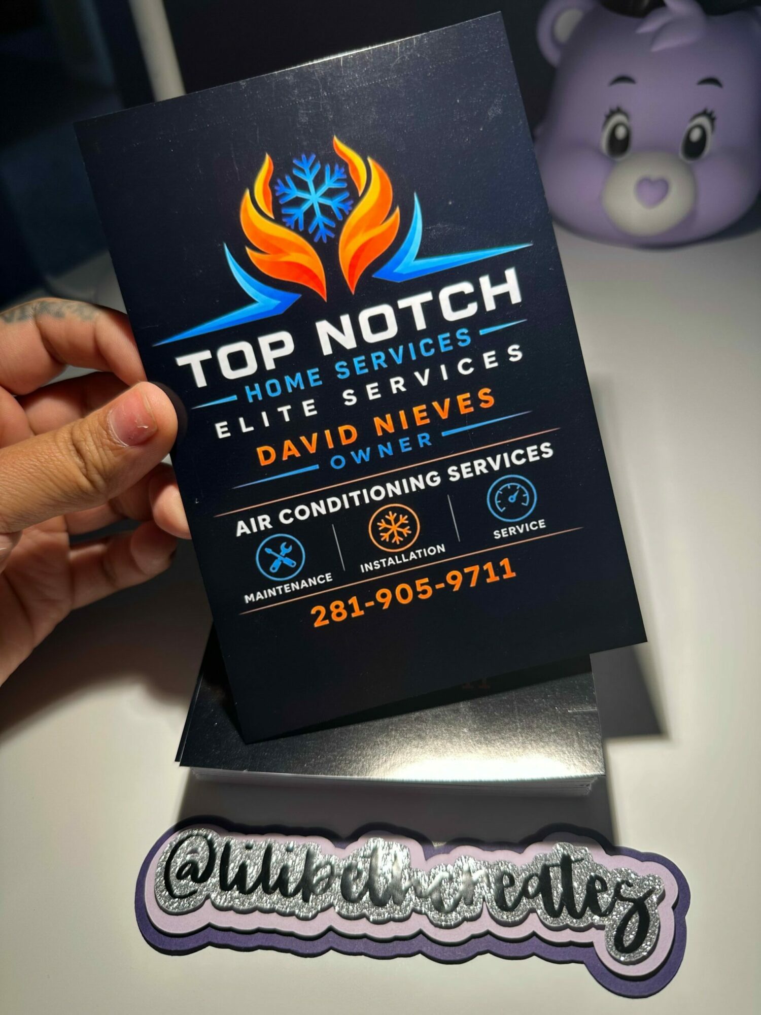 Person holding a business card with a logo and contact information, with a purple toy in the background, on a white surface.