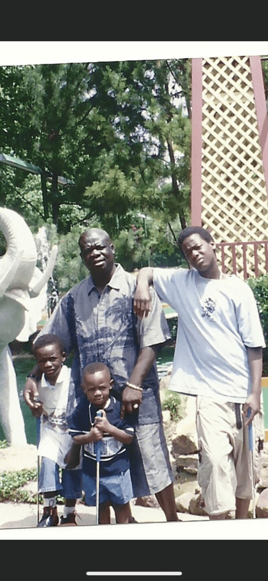 Group of five people standing outdoors with trees and a lattice structure in background.
