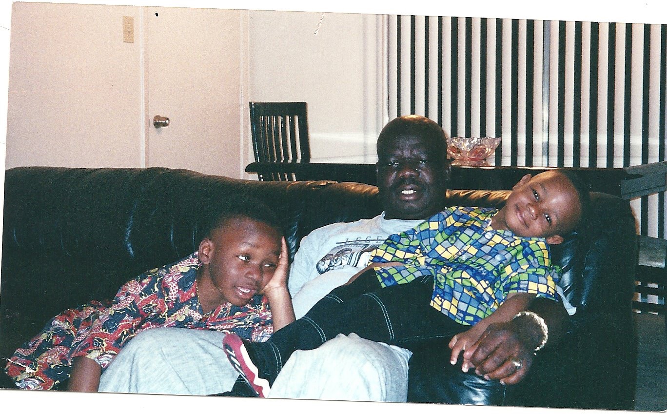 Three people sitting on a couch, two children and an adult, in a living room with a dining table in the background.