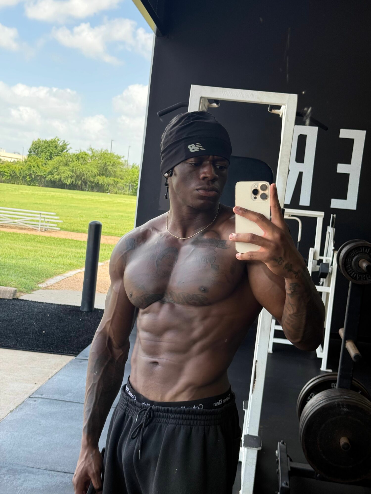Man taking a mirror selfie at the gym, showing his muscular upper body, outdoors background with trees and sky.