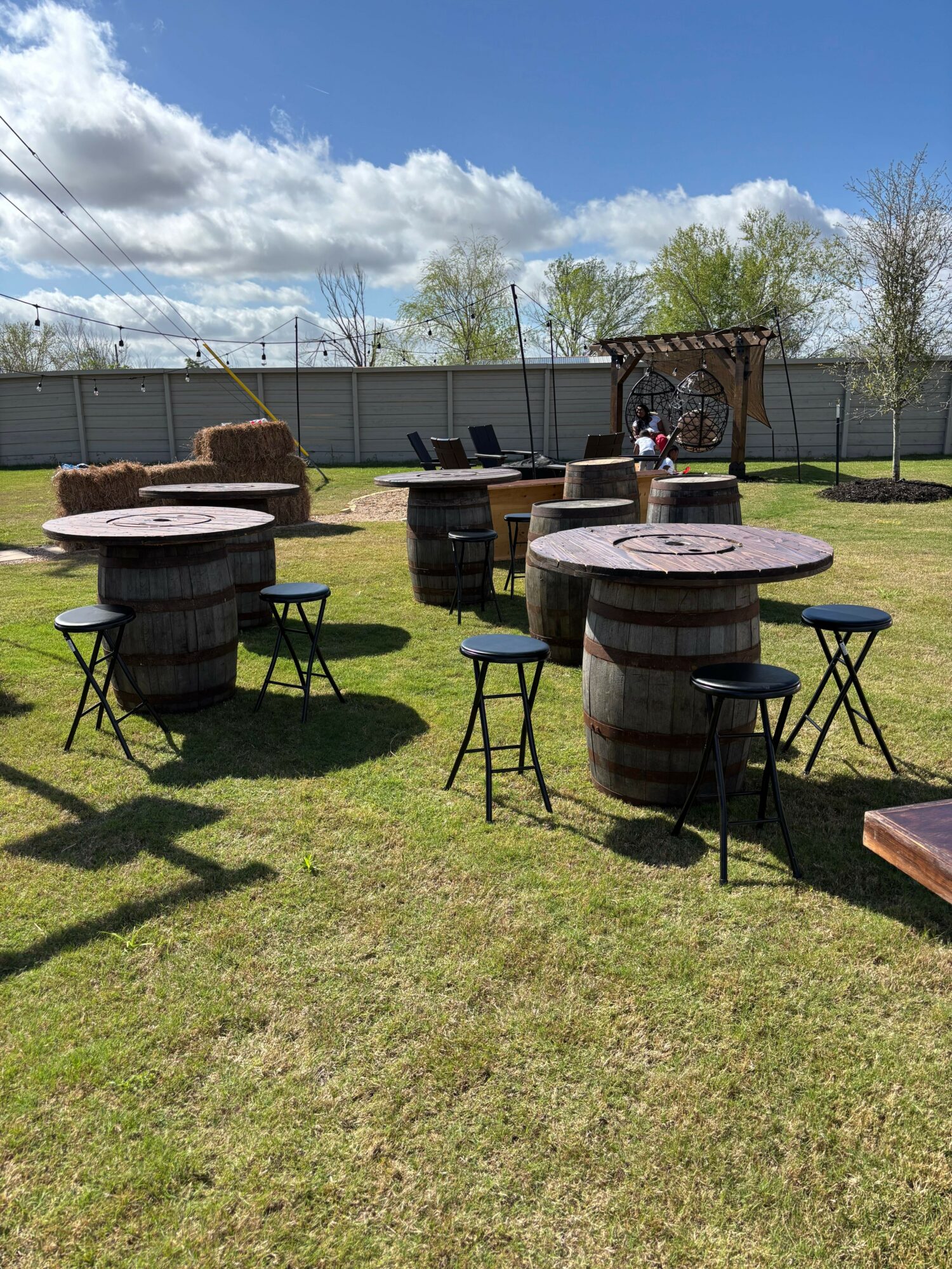 Outdoor scene with tables and chairs on grass, shadows cast on ground, fence and trees in background, cloudy sky.