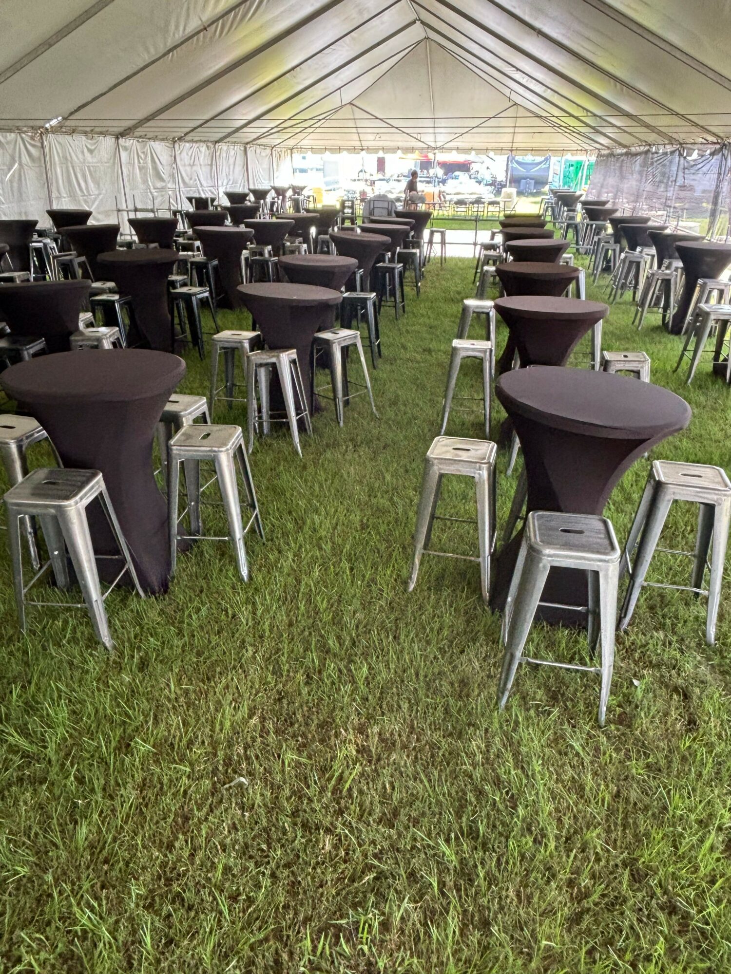 Outdoor seating area with black chairs and small tables on grass, under a canopy, with a building in the background.