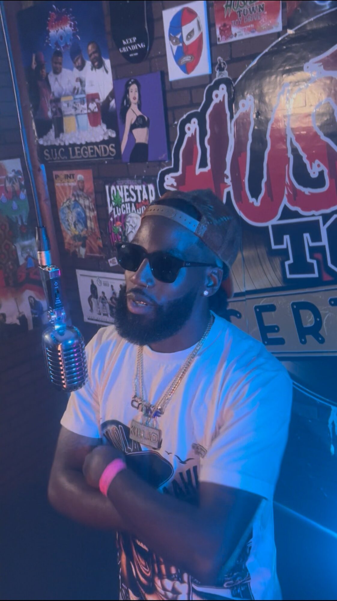 Man with sunglasses and beard stands with arms crossed in a colorful, poster-filled room.