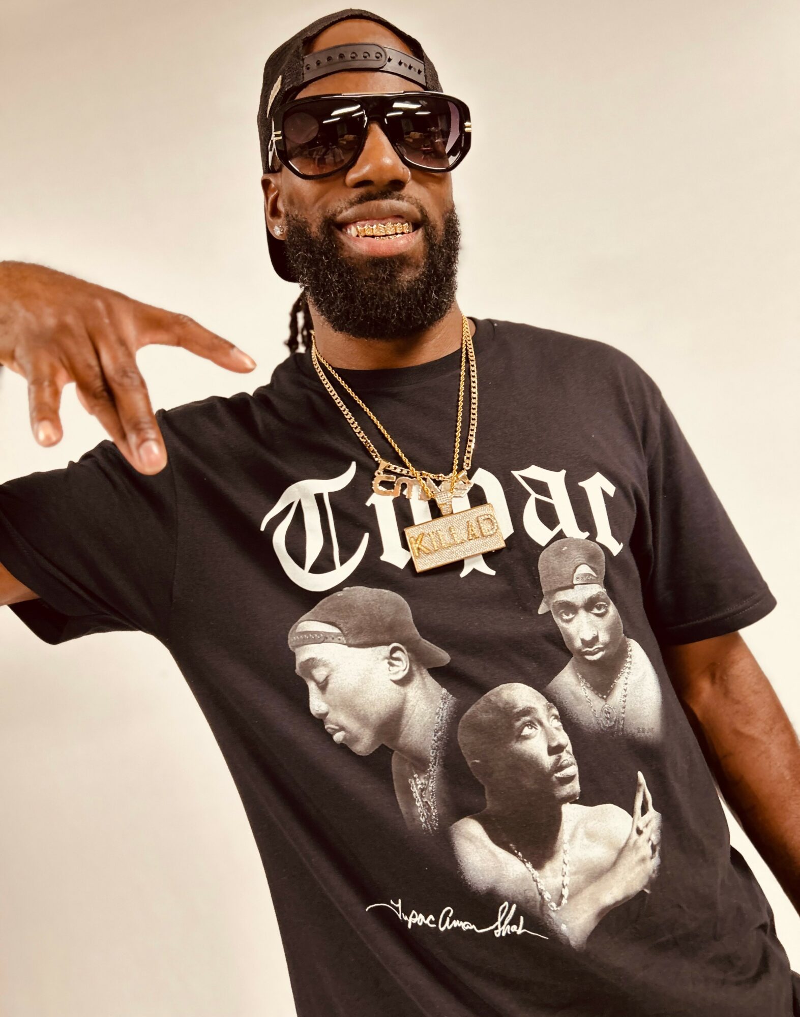 Man with sunglasses, beard, and cap smiling, wearing a black T-shirt with black-and-white portraits and text, pointing to the side.