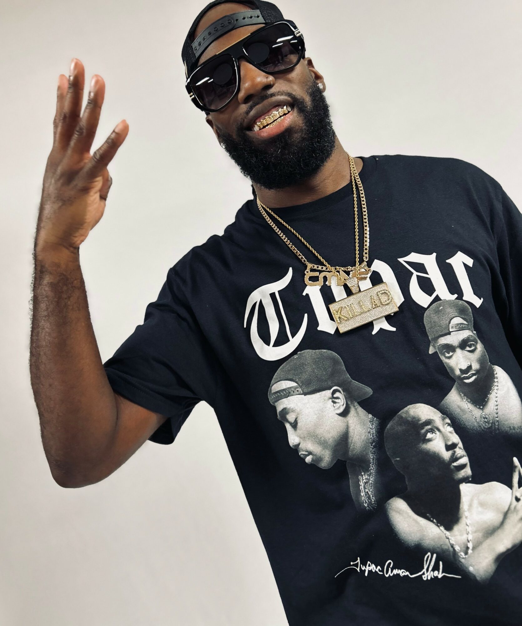 Man with sunglasses, beard, and cap, wearing a black T-shirt with a graphic of three people, smiling and making a hand gesture.