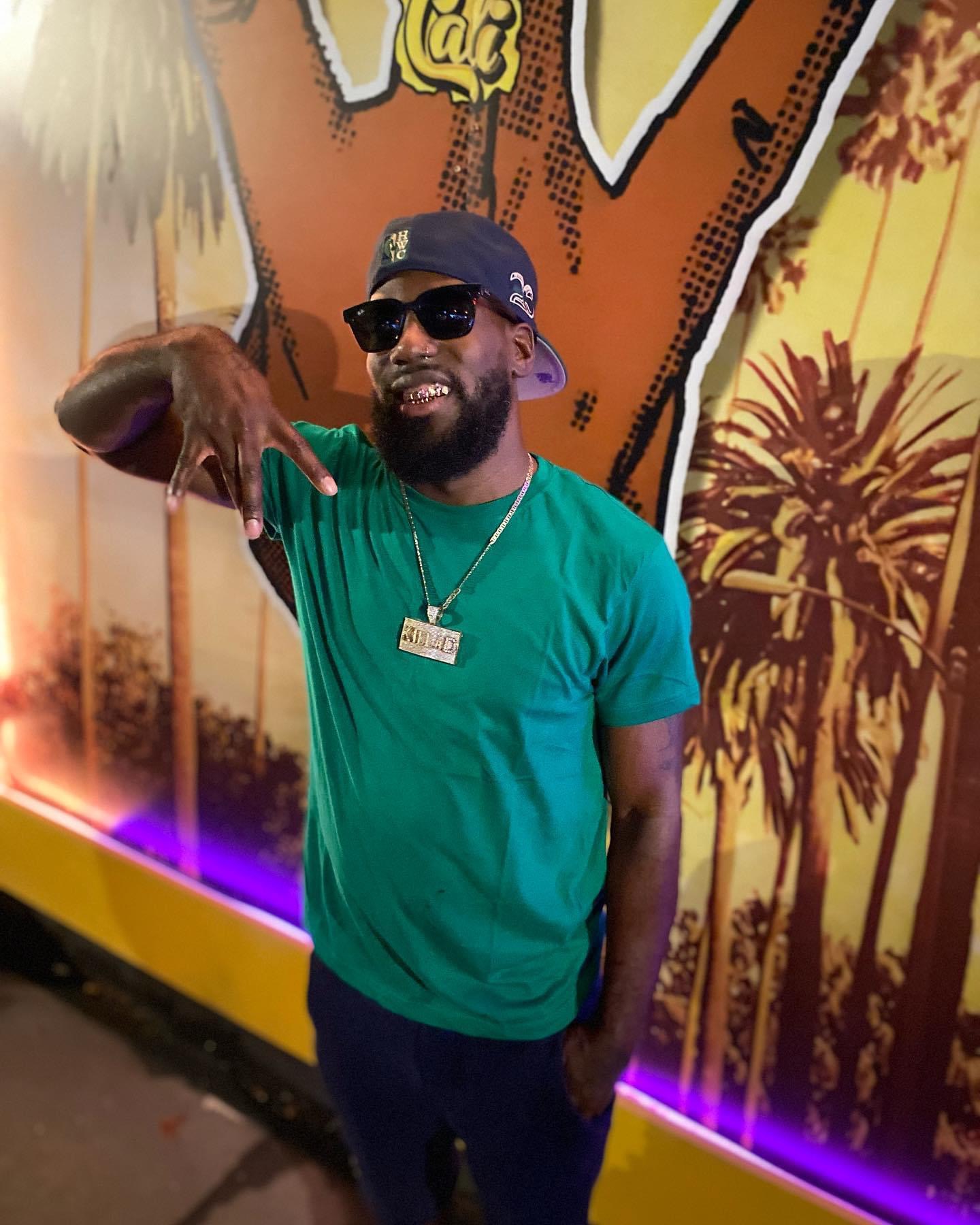 Man with sunglasses, beard, and cap smiling, pointing, standing in front of colorful mural with trees and sunset, wearing a green shirt.