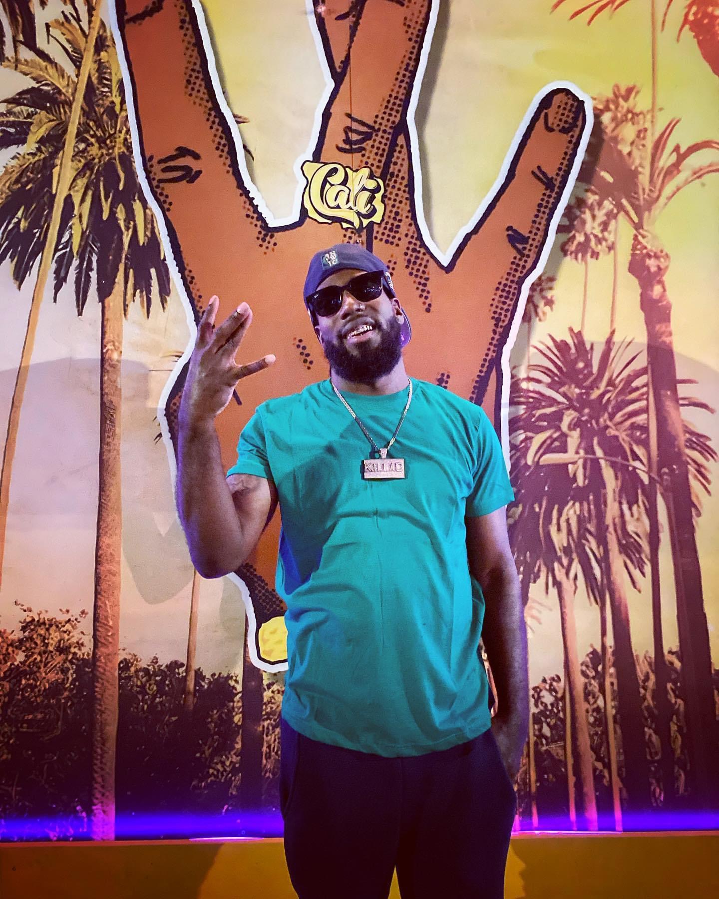 Man wearing sunglasses and a turquoise shirt making a peace sign, standing in front of a colorful palm tree background with a large hand and a 'Cat' sign.