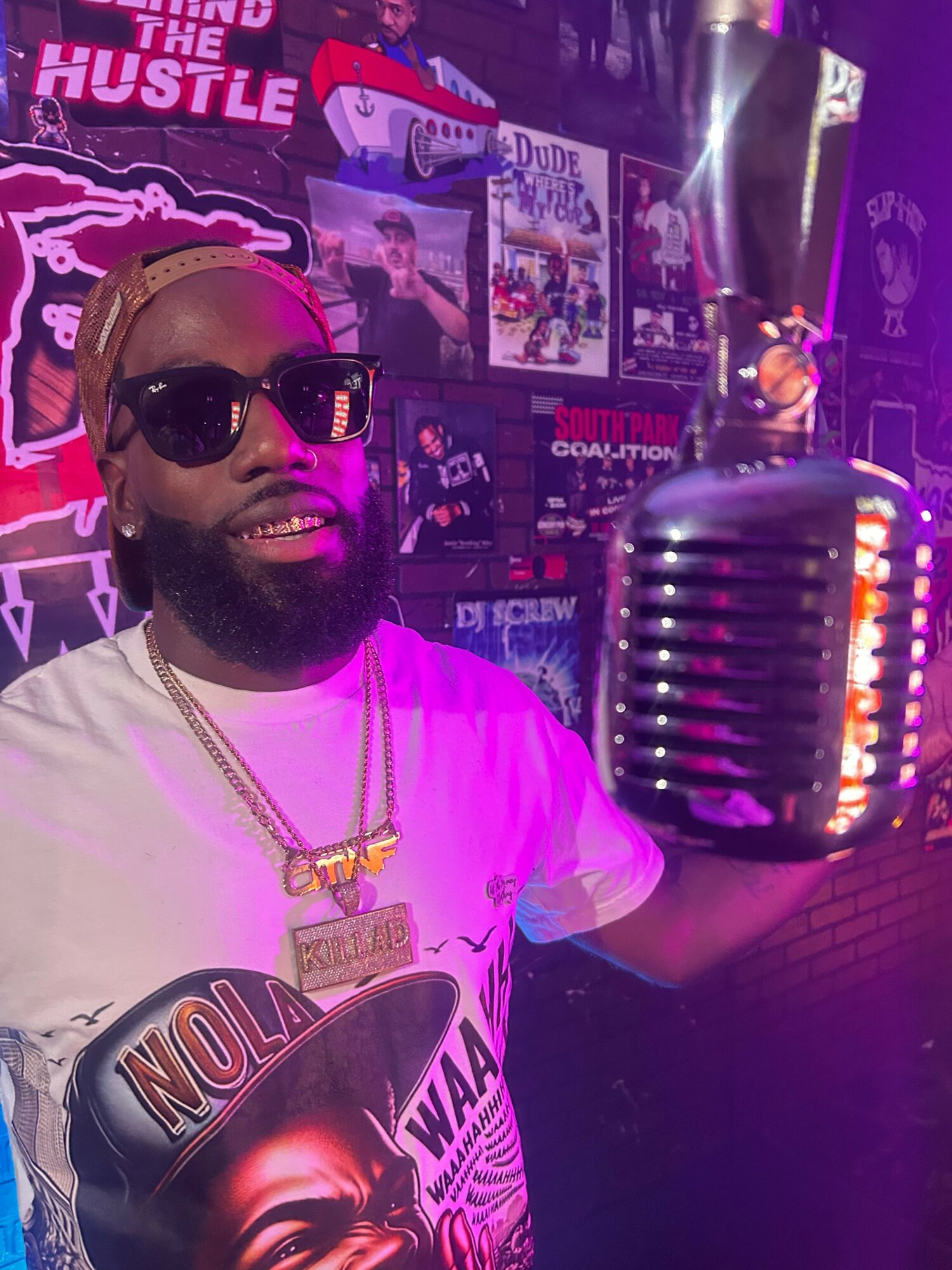 Person with sunglasses and gold chain in a colorful, music-themed environment, with a microphone and posters in the background.