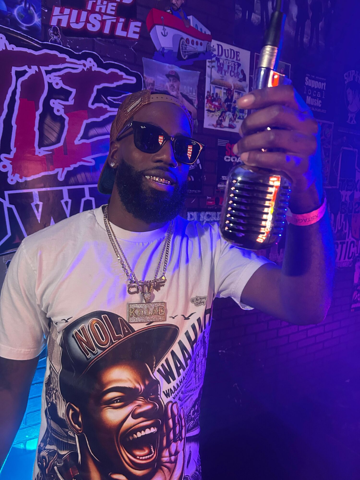 Person wearing sunglasses and a graphic t-shirt, holding a microphone, in a colorful, illuminated setting.