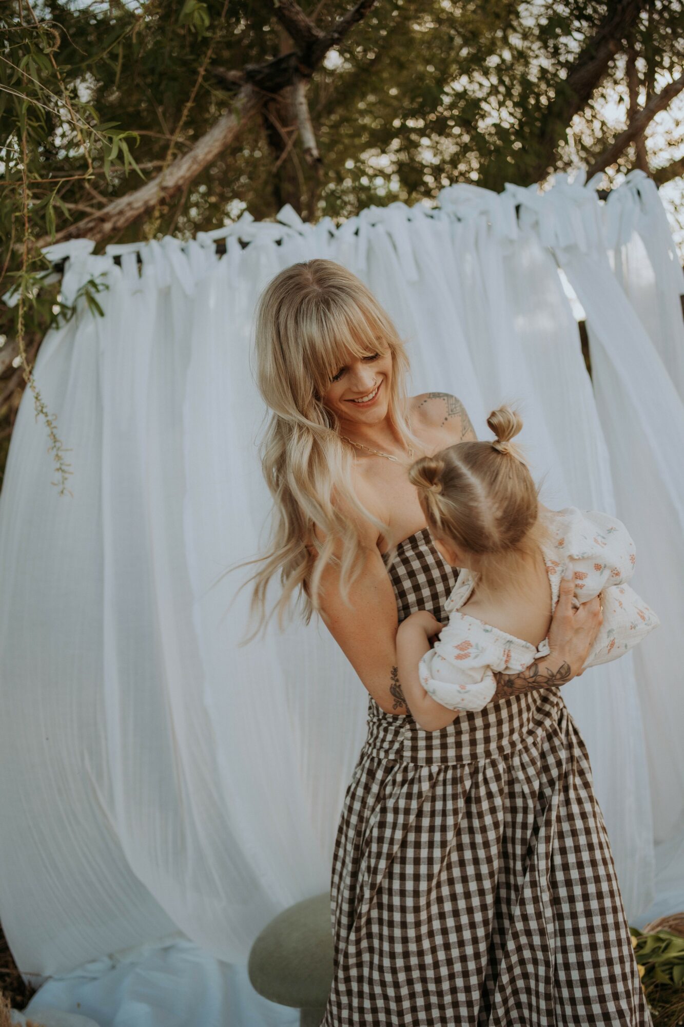 Woman holding two children outdoors with white fabric backdrop, trees, and greenery in background.