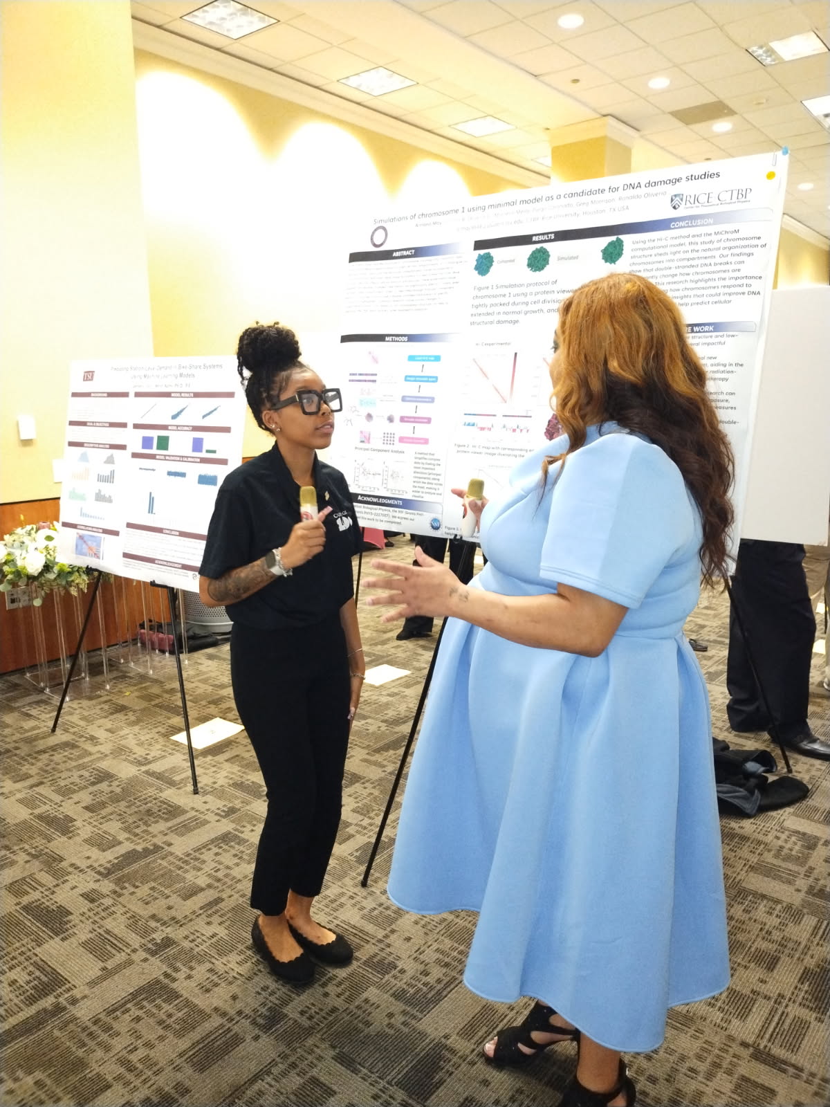 Two women are talking in front of research posters at an indoor conference, one holding a microphone and the other gesturing.