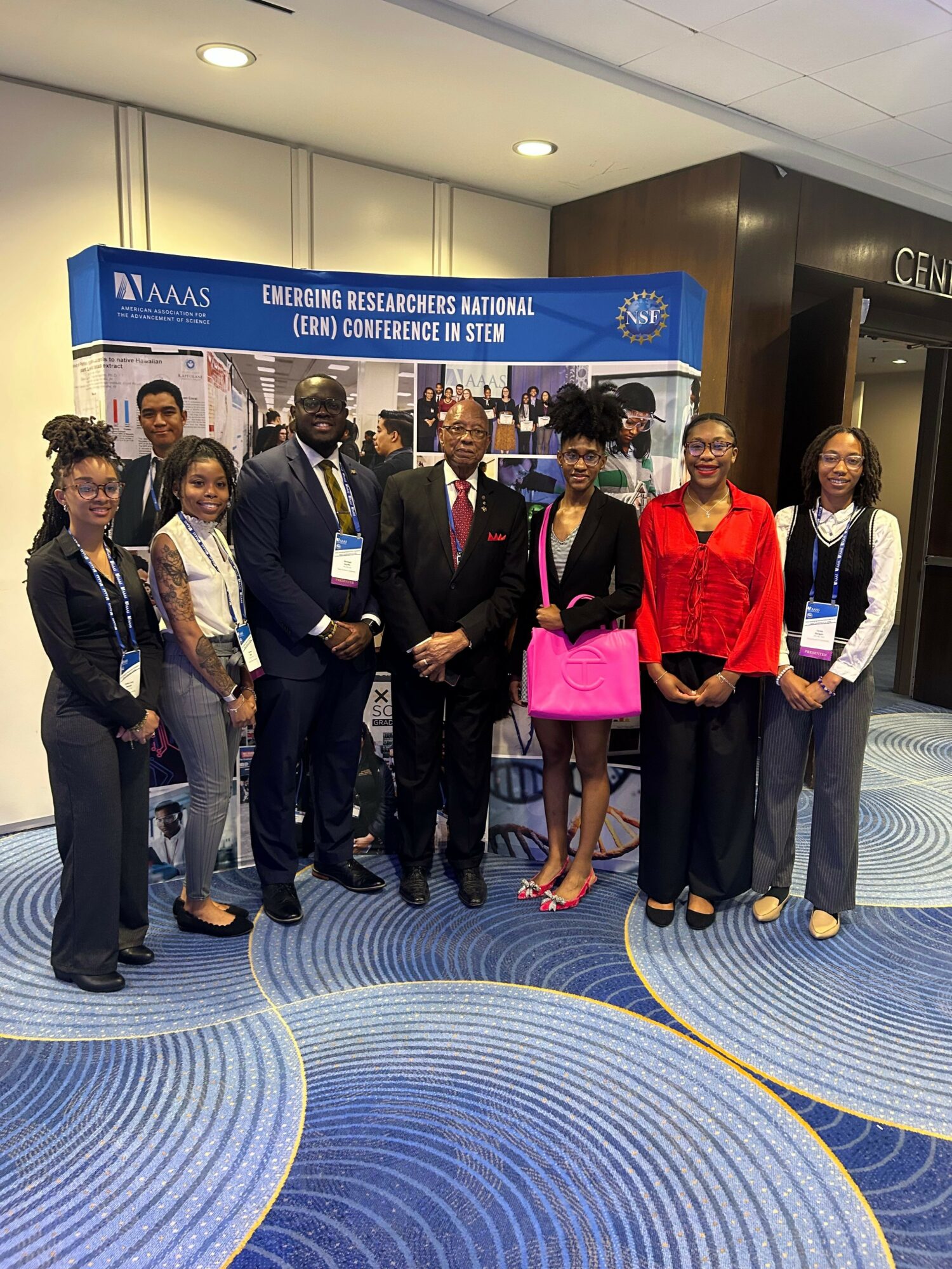 Group of seven people standing in front of a blue banner at an indoor event, some holding bags and wearing formal attire.