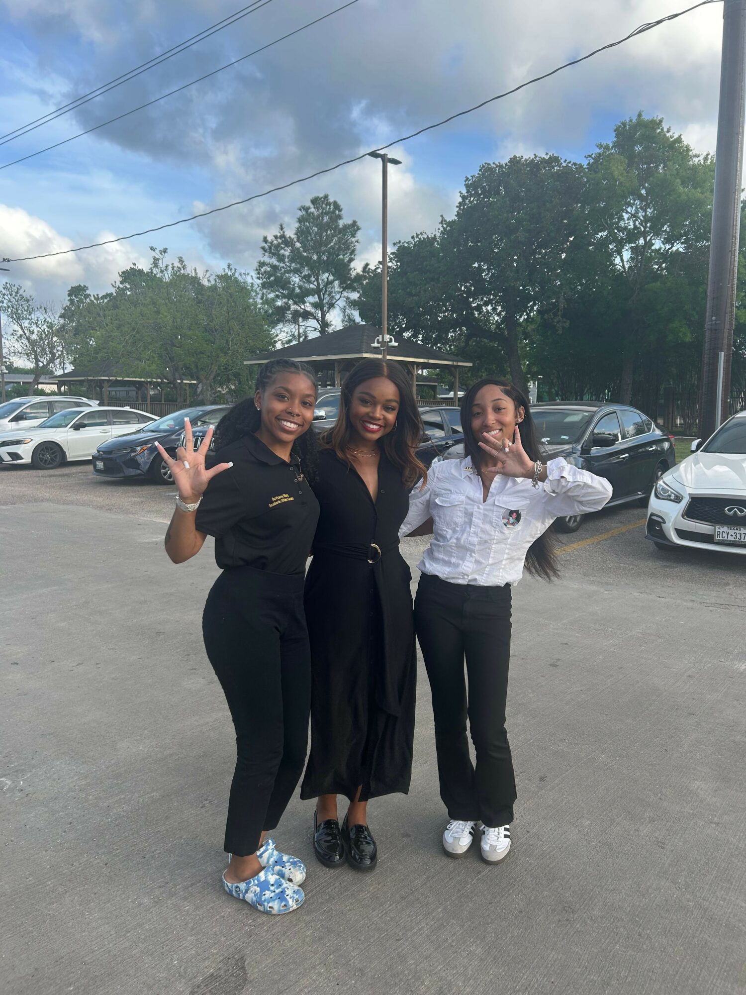 Three women standing outdoors in a parking lot, with cars and trees in the background, smiling and making peace signs.