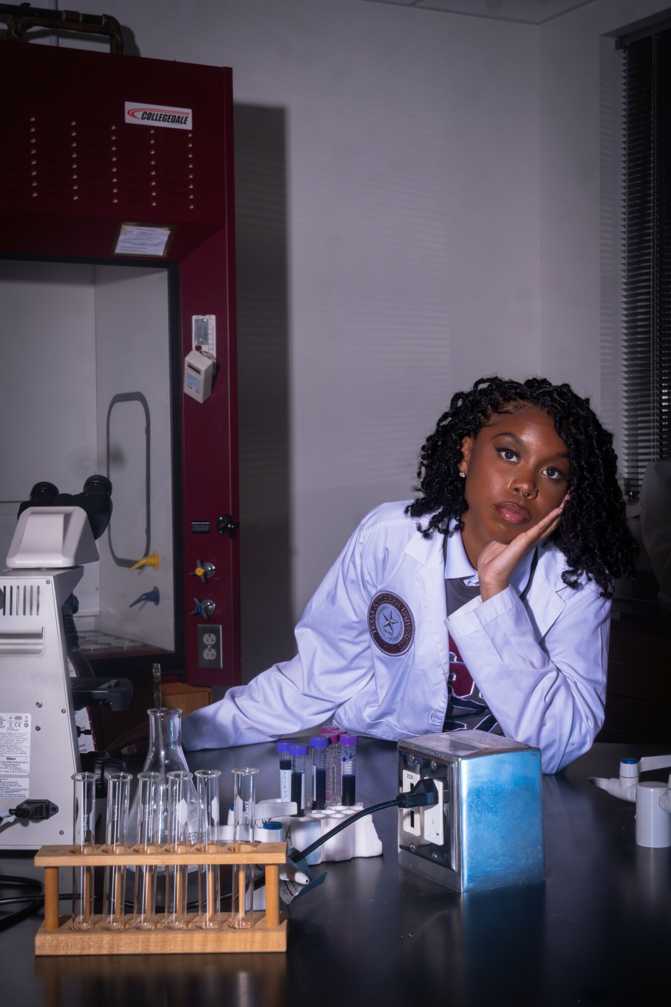 Female scientist in a lab coat resting her head on her hand at a laboratory bench with test tubes and equipment.