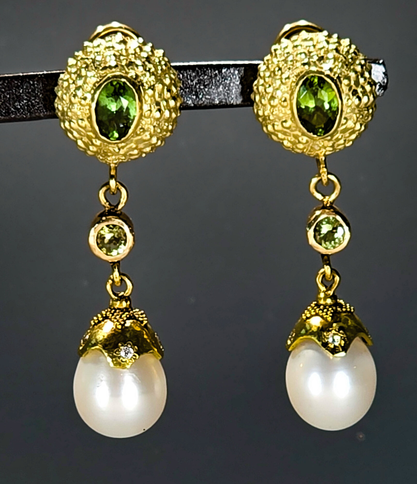 Gold earrings with green gemstones and pearl drops, hanging from a black display. Bright, detailed jewelry design.