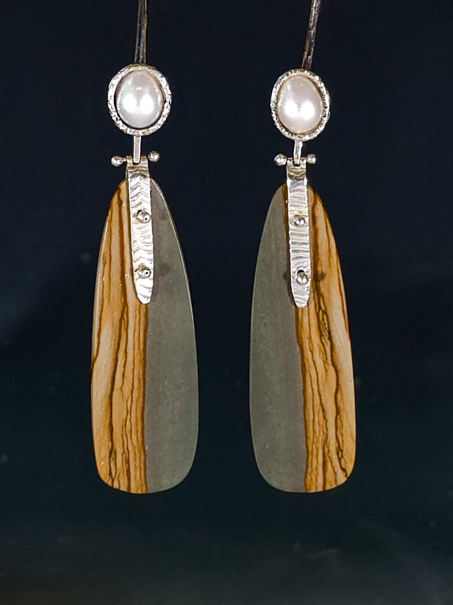 Pair of earrings with pearl tops, wooden and metal elongated pendants, and decorative silver accents, hanging against a dark background.