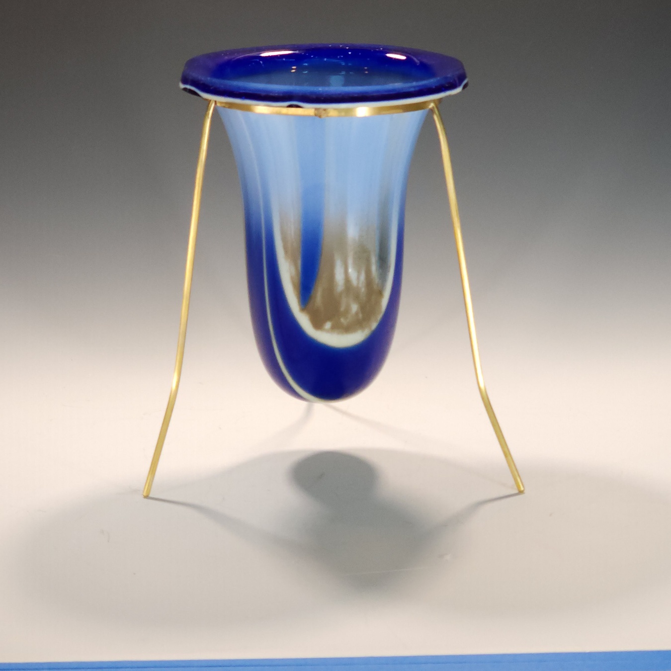 Blue glass vessel with gold-colored legs, standing on a white surface, casting a shadow, against a gray background.