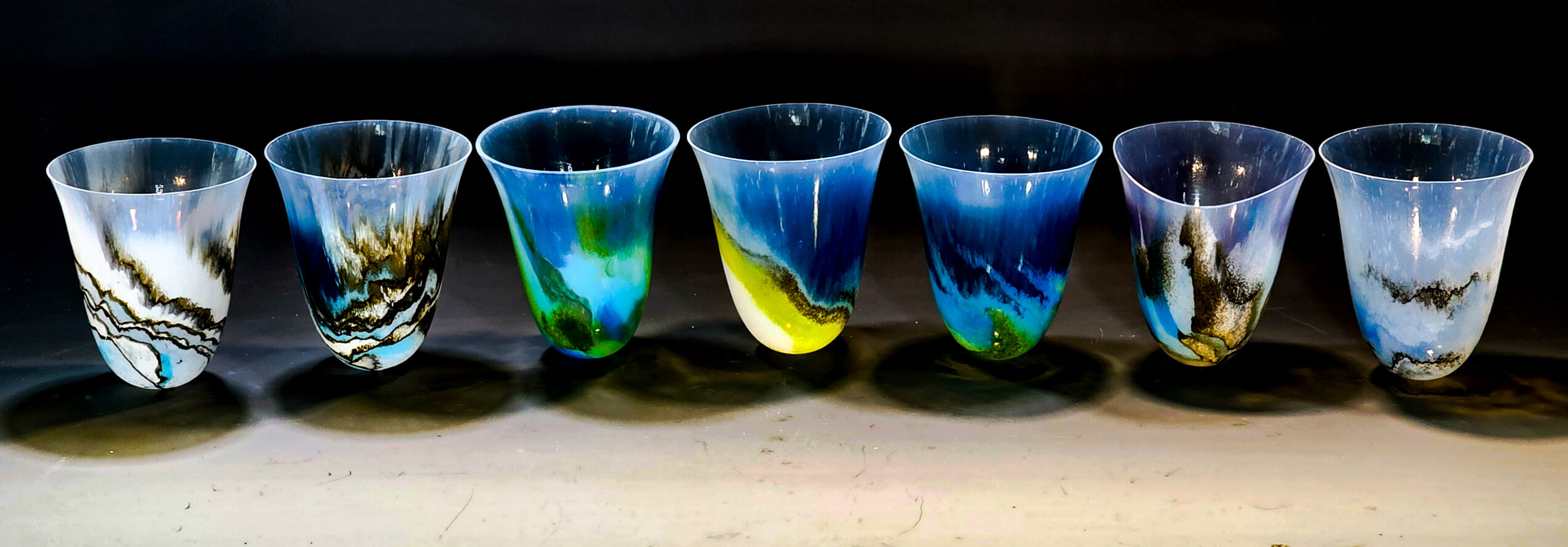 Seven glass cups with colorful abstract patterns, arranged in a row on a surface.
