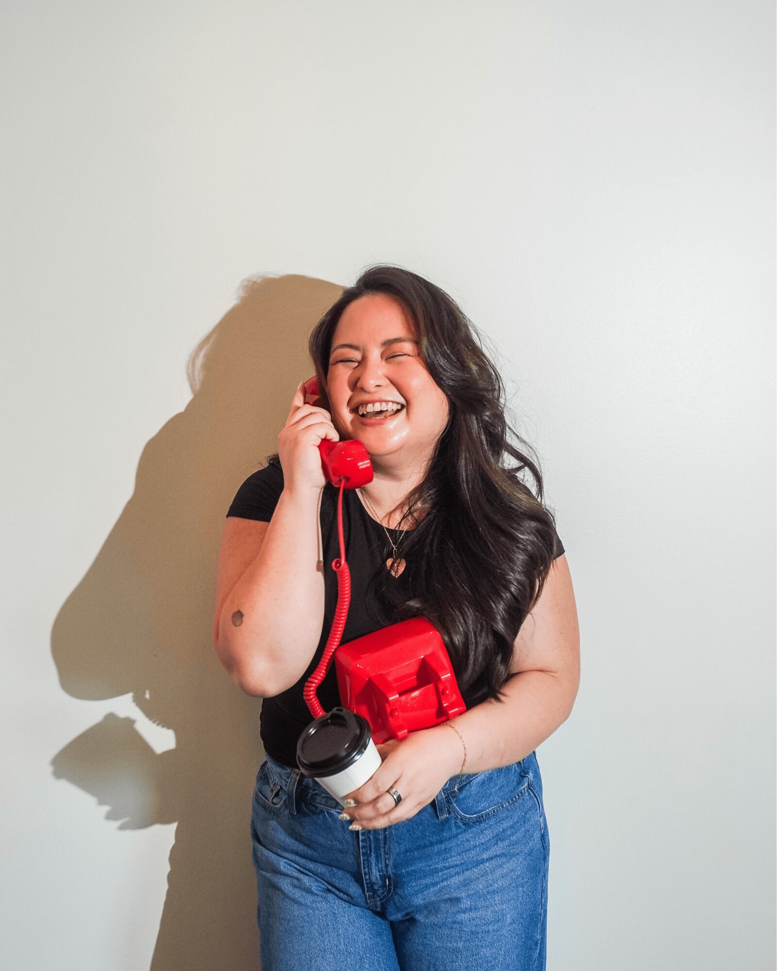 Young woman with long dark hair smiling while talking on a red vintage telephone, holding a camera, standing against a plain wall.