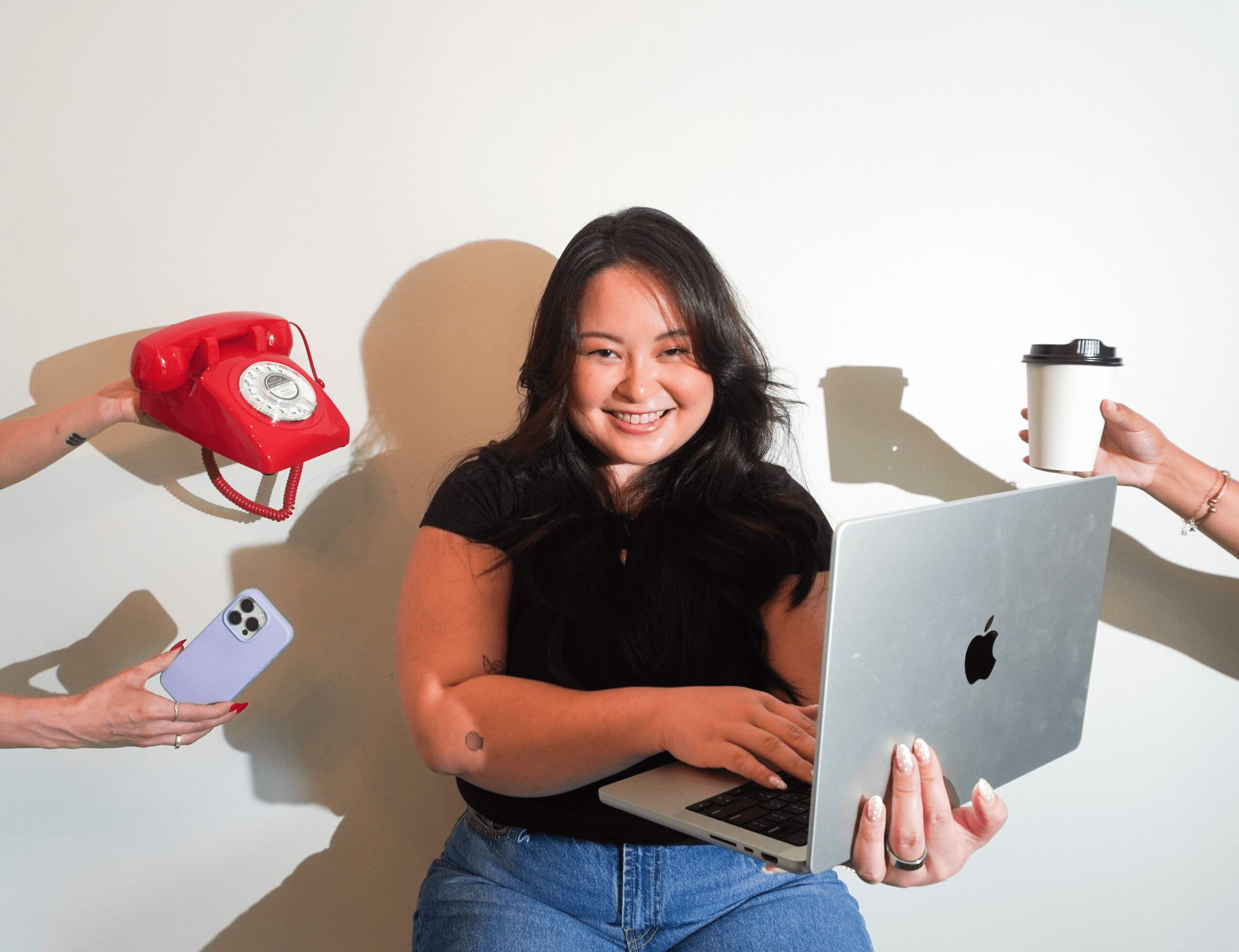 Young woman smiling while holding a laptop, with a red rotary phone and a coffee cup on either side.