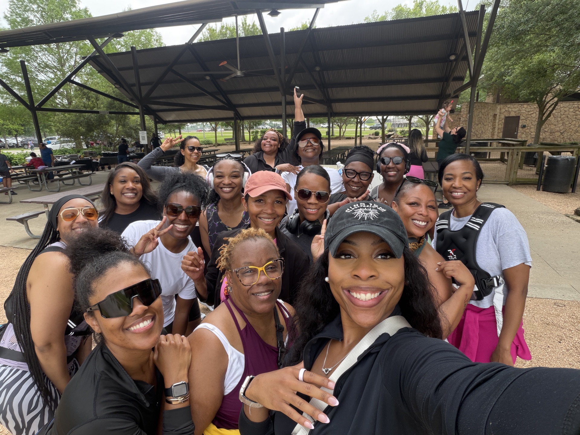 Group of smiling women and girls outdoors, some wearing sunglasses and hats, under a canopy, posing for a selfie.