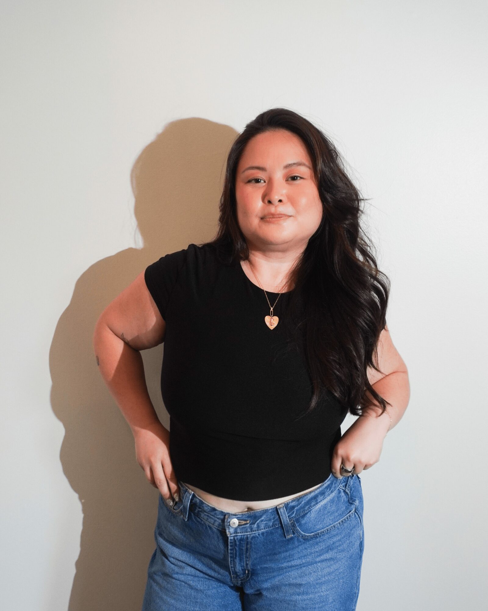Young woman with long dark hair wearing a black top and blue jeans, standing against a plain white background.