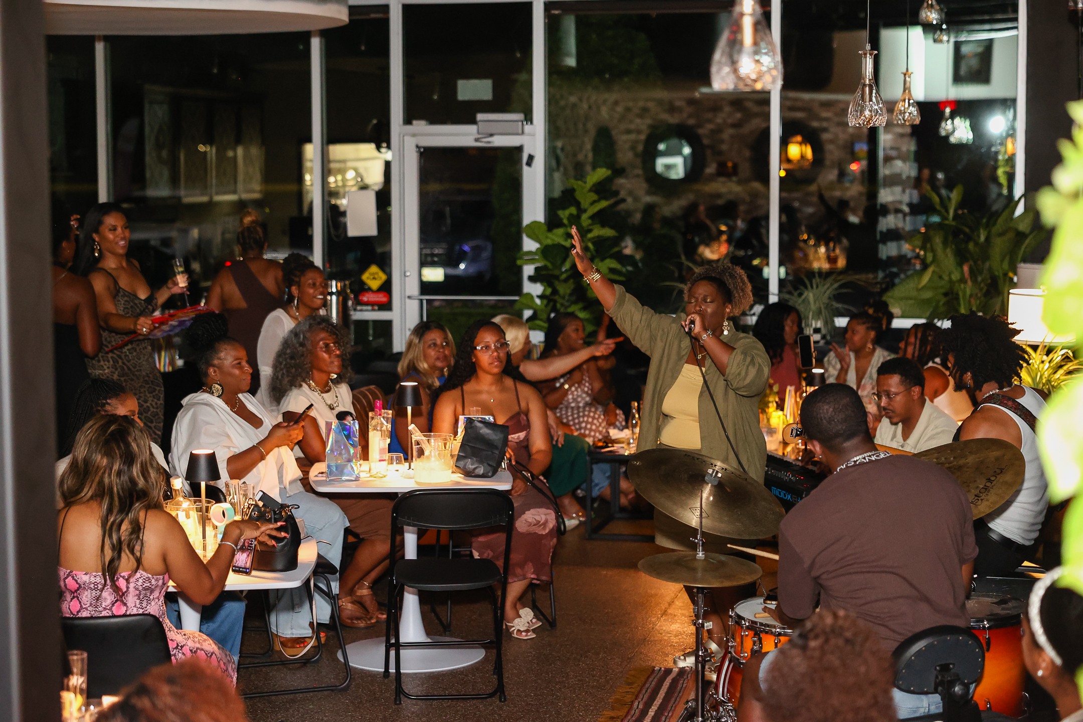 Group of people gathered around tables in a restaurant, some singing and others listening, with warm lighting and decor.