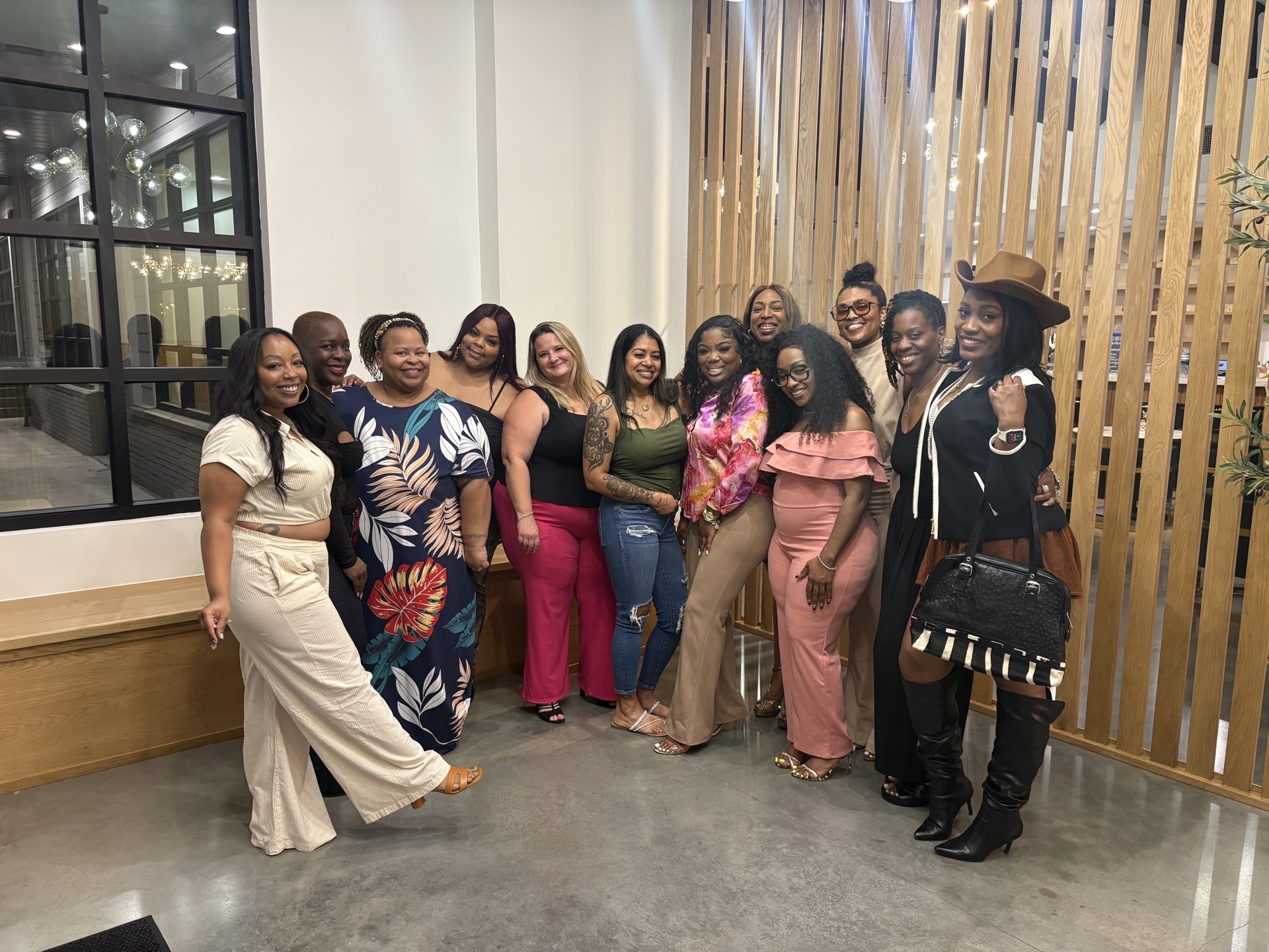 Group of women standing indoors, smiling, with wooden wall and large window in background.