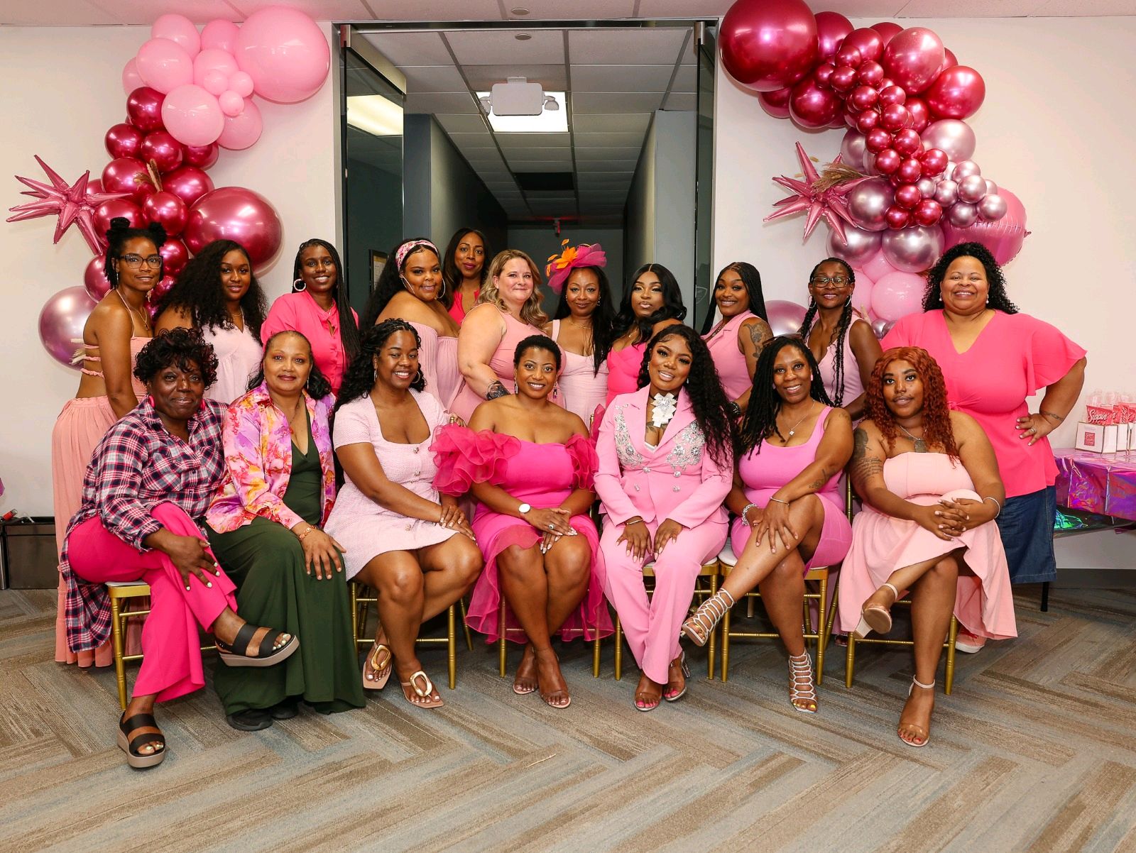 Group of women in pink and purple outfits sitting and standing in a decorated room with pink balloons.