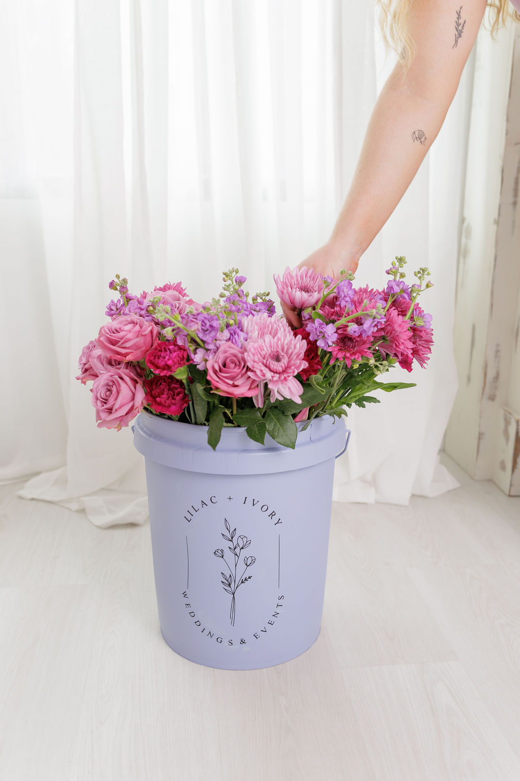 Person's arm reaching into a large purple flower vase with pink and purple flowers, in a bright room.